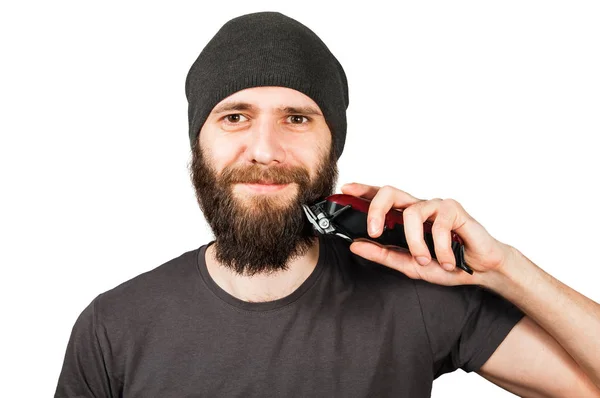 Jonge bebaarde man met hoed houdt Clipper vast en snijdt zijn baard door. Geïsoleerd op witte achtergrond — Stockfoto