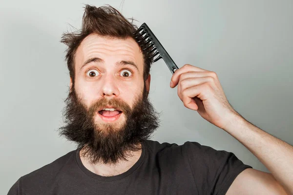 Joven barbudo sobrecrecido hombre sosteniendo peine y peinándose el pelo. Primer plano retrato sobre fondo gris —  Fotos de Stock