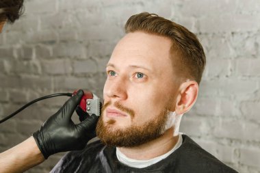 Eldiven Berber el saç kesmek ve bir tuğla duvar arka plan üzerinde yetişkin gihger sakallı adam tıraş. Bir adamın yakın portresi