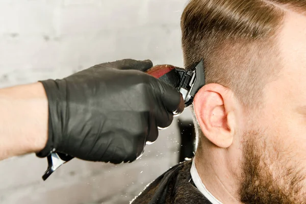 Barbeiro mão em luvas cortar o cabelo e barba adulto gihger barbudo homem em um fundo de parede de tijolo. Close up retrato de um cara — Fotografia de Stock
