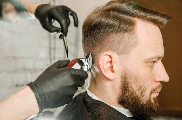 Friseurhand in Handschuhen schneidet Haare und rasiert erwachsenen bärtigen Mann auf Backsteinmauer Hintergrund. Nahaufnahme Porträt eines Mannes — Stockfoto