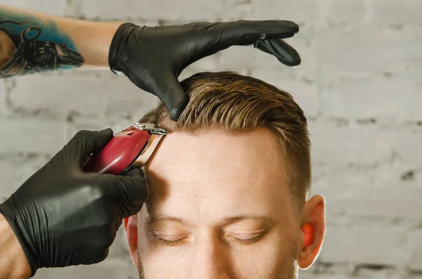 Friseurhand in Handschuhen schneidet Haare und rasiert erwachsenen bärtigen Mann auf Backsteinmauer Hintergrund. Nahaufnahme Porträt eines Mannes — Stockfoto