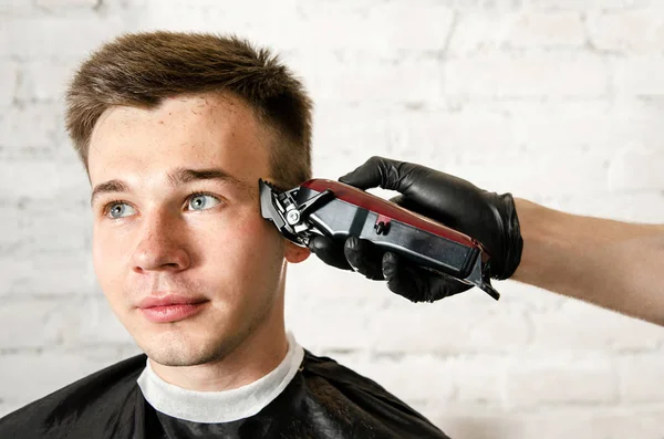 Friseurhand in Handschuhen schneidet Haare und rasiert jungen Mann vor Backsteinmauer-Hintergrund. Nahaufnahme Porträt eines Mannes — Stockfoto