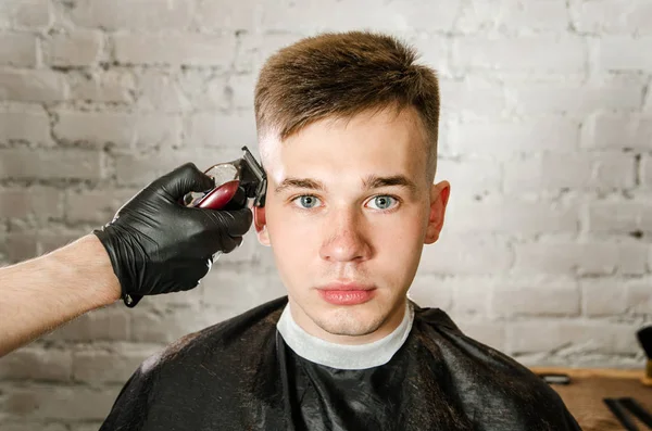 A mão de barbeiro em luvas corta o cabelo e rapa o jovem em um fundo de parede de tijolo. Close up retrato de um cara — Fotografia de Stock