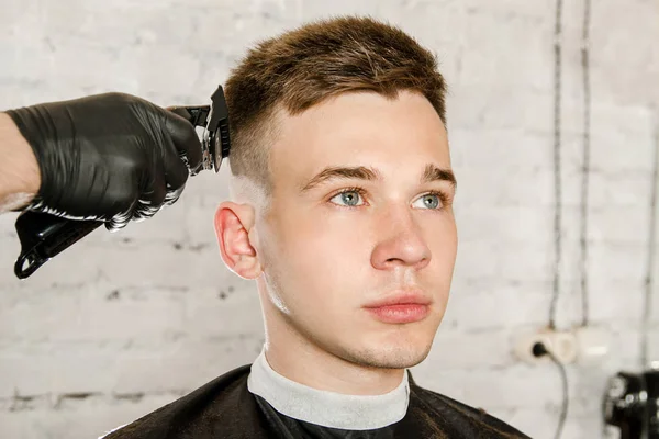 Friseurhand in Handschuhen schneidet Haare und rasiert jungen Mann vor Backsteinmauer-Hintergrund. Nahaufnahme Porträt eines Mannes — Stockfoto