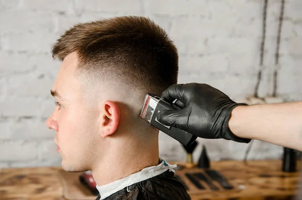 Barber hand i handskar klippa hår och rakar ung man på en tegelvägg bakgrund. Närbild porträtt av en kille — Stockfoto