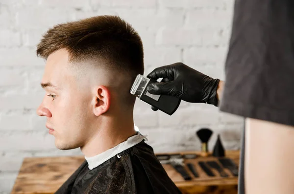 Friseurhand in Handschuhen schneidet Haare und rasiert jungen Mann vor Backsteinmauer-Hintergrund. Nahaufnahme Porträt eines Mannes — Stockfoto