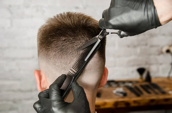 A mão de barbeiro em luvas corta o cabelo e rapa o jovem em um fundo de parede de tijolo. Close up side portrait de um cara, vista traseira — Fotografia de Stock
