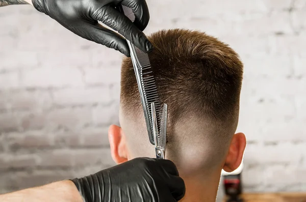 Barber hand in gloves cut hair and shaves young man on a brick wall background. Close up side portrait of a guy, back view