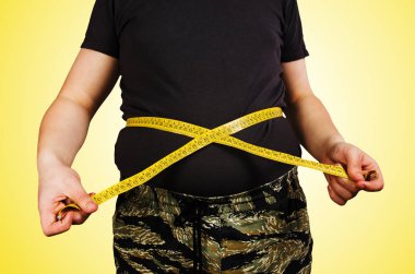 Body of young man with excess weight, keep belly fat in a T shirt, measure itself with a measuring tape on yellow background. clipart