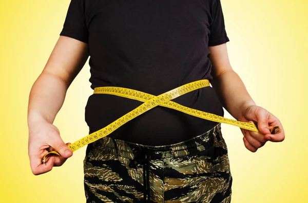 Szerve fiatal férfi a túlsúly, tartsa hasa zsírt egy póló, mérjük meg magát egy mérőszalag sárga háttér. — Stock Fotó