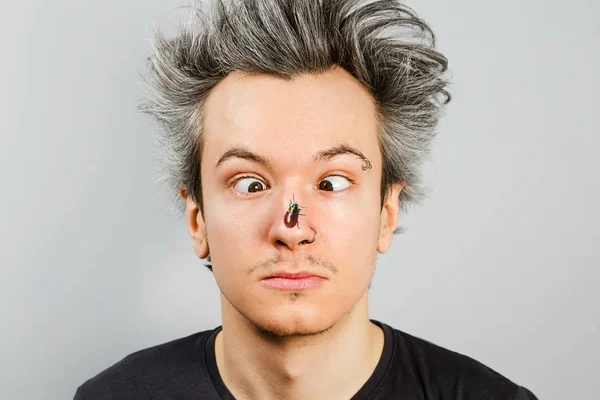 Unrasierter freak schielender junger Kerl mit Piercings im Gesicht, mit einem Käfer auf der Nase, auf grauem Hintergrund. — Stockfoto