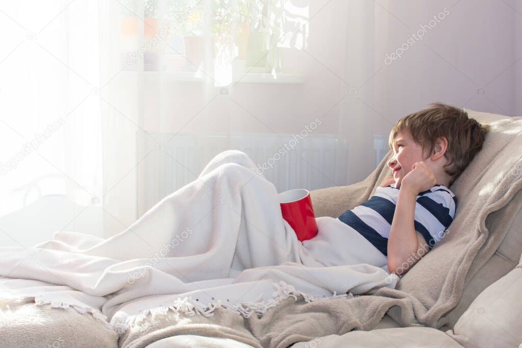 Boy is lying in bed under a blanket on a Sunny morning near window.