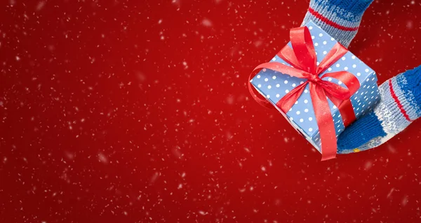 Manos en manoplas está sosteniendo una caja de regalo con cinta en el fondo rojo con nieve. Feliz tarjeta de felicitación de Navidad. —  Fotos de Stock