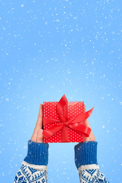 Manos está sosteniendo una caja de regalo roja con cinta en fondo azul con nieve. Feliz tarjeta de Navidad. — Foto de Stock