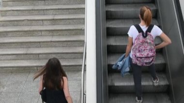 Sırt çantalı iki genç kadın bir metro istasyonunda merdivenleri ve yürüyen merdivenleri kullanarak yukarı çıkıyorlar.