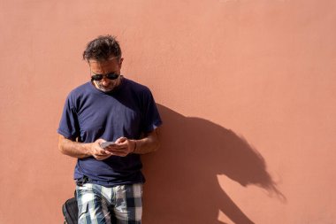 Yakışıklı adam, güneşli bir günde güneş gözlüğü takarak cep telefonunu kullanarak boş bir arkaplanda poz veriyor.