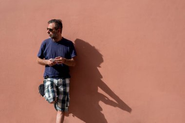 Yakışıklı adam, güneşli bir günde güneş gözlüğü takarak cep telefonunu kullanarak boş bir arkaplanda poz veriyor.