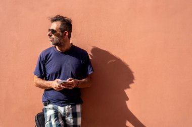 Yakışıklı adam, güneşli bir günde güneş gözlüğü takarak cep telefonunu kullanarak boş bir arkaplanda poz veriyor.