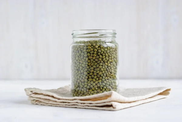 Glass storage jar full of dried green mung beans on a kitchen cloth over a white background viewed from the side