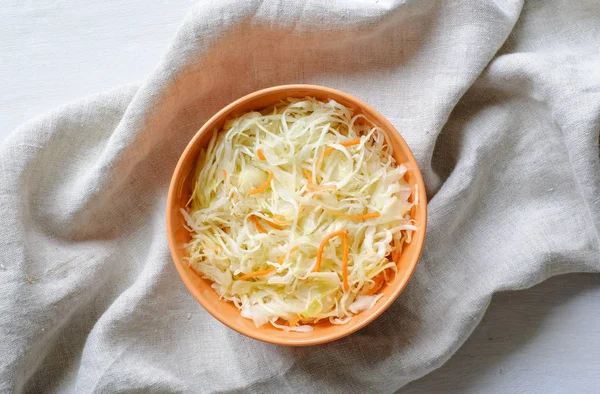 Top view of orange bowl of sauerkraut with chopped cabbage and carrot on grey towel sitting on white table surface
