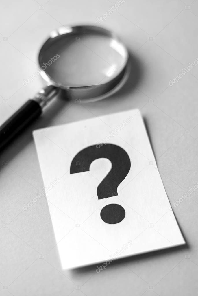 Magnifying glass on grey surface, next to big black question mark printed on white sheet of paper. Search for answers concept