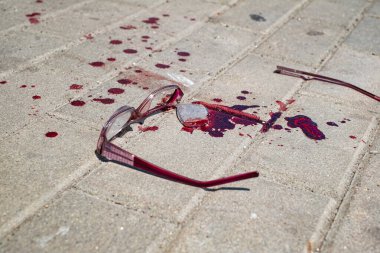 A pair of red-framed eyeglasses lies broken on a light gray paved surface, A significant amount of  blood is splattered and pooled around the eyeglasses