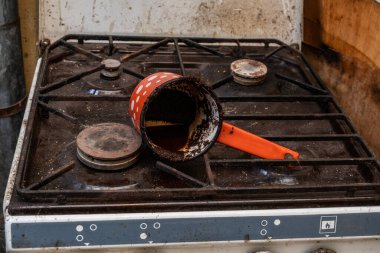 Paslanmış gaz sobası ve parlak turuncu saplı kahve dolu bir demlik. Tencere eğilmiş, içindekilerin bir kısmı ocağın üzerine dökülüyor. Sobanın ocakları kirli. dağınık bir mutfak ortamı