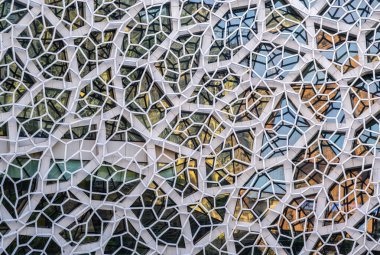a modern architectural facade featuring a complex, geometric pattern of white, hexagonal and irregularly-shaped frames. The frames enclose various glass panels, creating a mosaic-like effect.