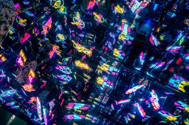 Neon ışıklı, yoğun bir şehir gece hayatı sahnesi. Sahne çeşitli tabelalarla, barlarla, kulüplerle ve dükkanlarla dolu. Atmosfer hareketli bir şehir gece hayatının özünü yakalıyor.