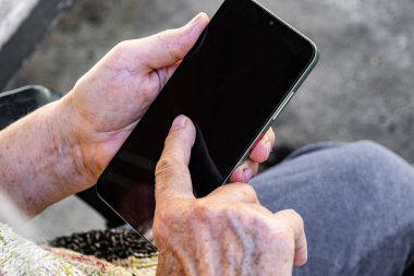 an elderly person with fair skin holding a black smartphone. The person wears a patterned sweater with shades of beige, green, and yellow. The background is blurred, showing a grayish pavement