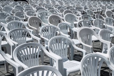 Photograph of an outdoor seating arrangement featuring numerous white plastic chairs and tables arranged in rows on a sunlit, cobblestone pavement