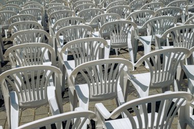Photograph of an outdoor seating arrangement featuring numerous white plastic chairs and tables arranged in rows on a sunlit, cobblestone pavement
