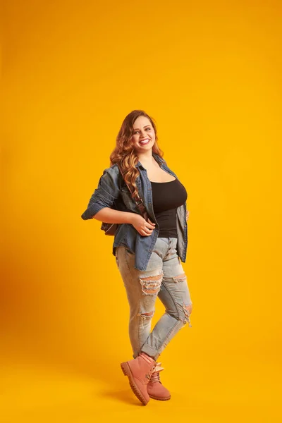 Candid curvy girl in denim jacket holding backpack and looking a — Stock Photo, Image