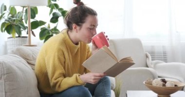 Kadın kitap okuyor ve evdeki kanepede kahve içiyor.