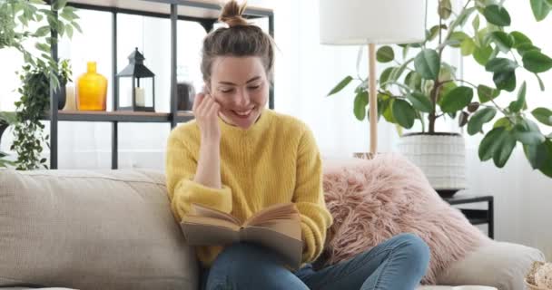 Γυναίκα διαβάζει ένα βιβλίο και χρησιμοποιεί το τηλέφωνο στον καναπέ στο σπίτι — Αρχείο Βίντεο