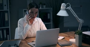 İş kadını cep telefonuyla konuşuyor ve gece ofisinde laptop kullanıyor.