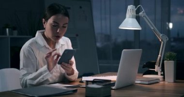 İş kadını telefonunu kullanıyor ve gece ofisinde dizüstü bilgisayarla çalışıyor.