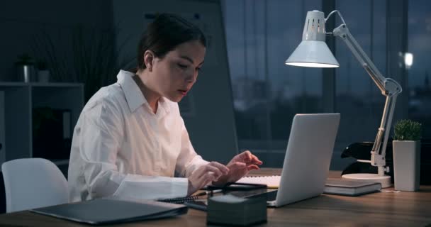 Businesswoman pracuje na notebooku a posílá hlasovou zprávu pomocí mobilního telefonu v noční kanceláři — Stock video