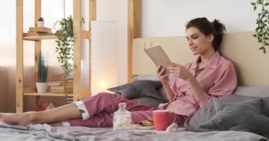 Kadın yatakta kahvaltı yaparken kitap okuyor.