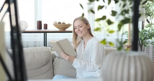 Mulher feliz lendo um livro no sofá em casa — Vídeo de Stock