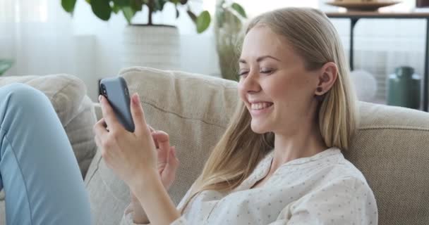 Счастливая женщина пользуется мобильным телефоном на диване дома — стоковое видео