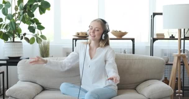 Радостная женщина наслаждается музыкой, используя наушники на диване — стоковое видео