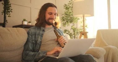 Adam dizüstü bilgisayar kullanıyor ve evde bira içiyor.