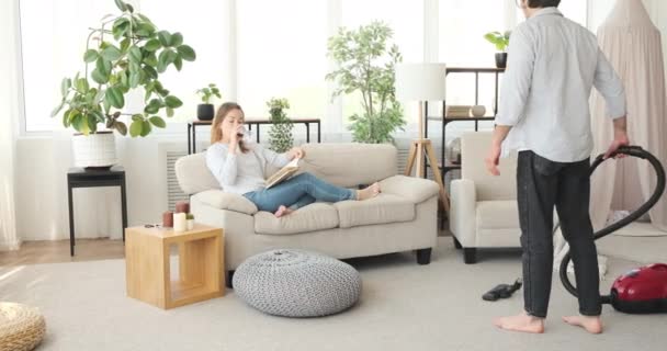 Vrouw drinkt wijn en leest boek terwijl man stofzuigt tapijt — Stockvideo