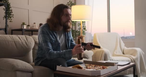 Чоловік насолоджується піцою та пивом, дивлячись телевізор вдома — стокове відео