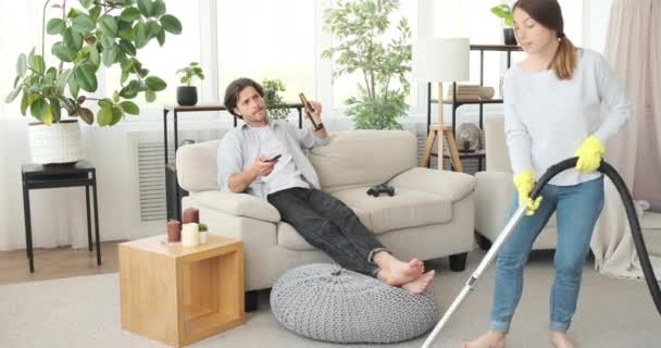 Mujer aspirando alfombras mientras su marido está bebiendo cerveza y viendo la televisión — Vídeo de stock