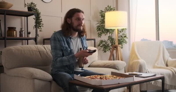 Человек, наслаждающийся пиццей и пивом, смотрит телевизор дома — стоковое видео