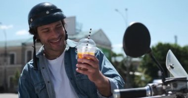 Adam şehirde motosiklette oturmuş meyve suyu içiyor.