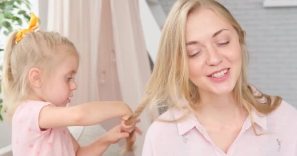 Маленькая девочка делает косы волос своей матери дома — стоковое видео